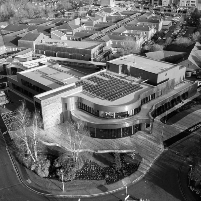 Christies Hospital – 500x500px blog image-01 Aerial view of a modern, curved building with surrounding roads and nearby residential buildings. Black and white photo.