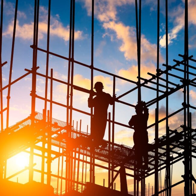 Two construction workers stand on scaffolding at sunset with a colorful sky in the background.